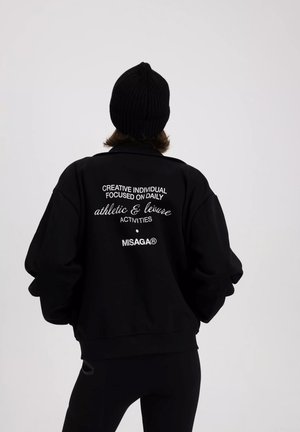 Zwart oversized sweatshirt met witte tekst op de achterkant, gecombineerd met een geribde zwarte beanie. Zacht materiaal en een relaxte pasvorm met verlaagde schouders.