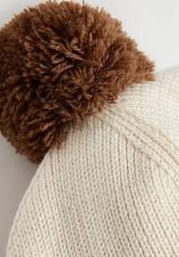 Strikket beanie med en cremefarvet krop og et struktureret mønster, toppet med en fluffy brun pompon. Blødt materiale til varme.