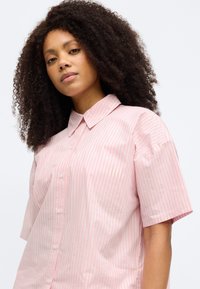 Ροζ ανοιχτό ριγέ κοντομάνικο πουκάμισο με λεπτές κάθετες ρίγες, κουμπιά μπροστά και μυτερό γιακά. Κατασκευασμένο από απαλό ύφασμα.