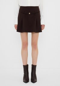 Jupe mini marron avec des plis, une taille ajustée et une boucle en or. Associée à des bottines marron élégantes. Texture de tissu lisse.