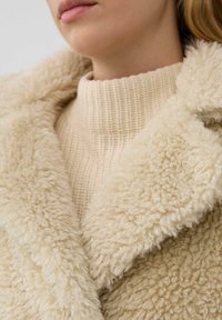 Close-up van een persoon met een beige, grof gebreid sweater en een pluizige, textuurvolle kraag van cremekleurige jas.
