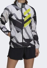 Letvægts vindjakke i hvid, sort og grå med et geometrisk mønster; har en høj krave og en markant gul grafisk accent.