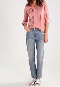 Blouse plissée rose avec un col en V, associée à un jean droit bleu clair et des sandales argentées à enfiler. Tissu lisse avec manches retroussées.