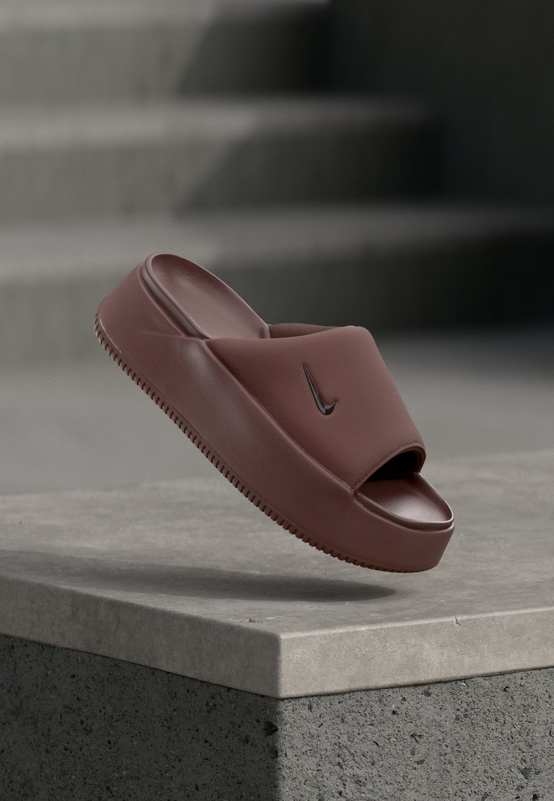 Rjava gumijasta natikača z odprtimi prsti, širšim sprednjim trakom in strukturo podplata. Na traku ima črn Nike logotip v obliki kljuka.