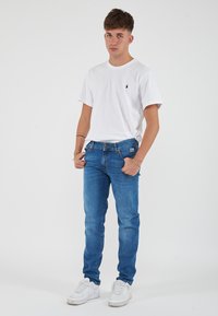 T-shirt bianco in cotone con maniche corte, jeans in denim blu e sneakers bianche. I jeans sono attillati, con una leggera scoloritura; il t-shirt presenta un piccolo logo.