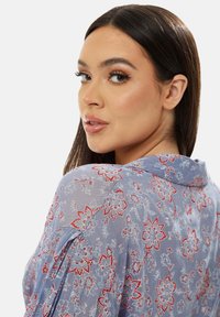 Light blue floral blouse with red and white patterns, featuring a collar and delicate, sheer fabric. The design includes pleated sleeve accents.