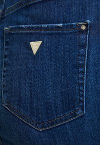 Mörkblå denimjeans med en triangelformad guld Guess-logotyp på bakfickan, dekorativ sömnad och subtila blekningdetaljer.