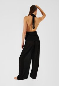 Achteraanzicht van een vrouw die een zwarte haltertop met strik en losse zwarte broek draagt. De stof is lichtgewicht en gestructureerd.