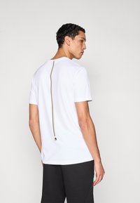 Biele tričko s krátkym rukávom, ktoré má výrazný čierny zips pozdĺž chrbta. Vyrobené z hladkej látky s voľným strihom.