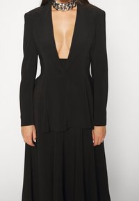 Černý blazer s hlubokým výstřihem do V, přiléhavým střihem, dlouhými rukávy a jemnými sklady na lemu. Nosený přes odpovídající černý top. Stříbrný řetízek.