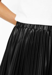 Jupe plissée noire en similicuir brillant, dotée d'une taille élastique et de côtes verticales, vue en gros plan de la taille vers le bas.