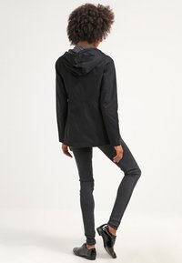 Person med krulligt hår som bär en svart huvdjacka, mörka tajta jeans och svarta glänsande skor, stående med ett ben något lyft.