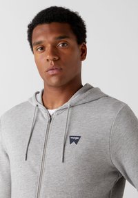 Szara bluza z kapturem na zamek, z regulowanym kapturem, z granatowym logo na lewej piersi, wykonana z miękkiej, teksturowanej bawełny.