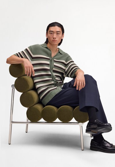 Giovane uomo con maglia a righe verdi a maglia e pantaloni blu navy seduto a gambe incrociate su una moderna poltrona cilindrica verde oliva.