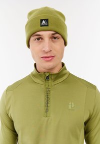Jonge man in een bijpassende olijfgroene beanie en een ritsjack waarop het logo "PROTEST" staat, tegen een witte achtergrond.