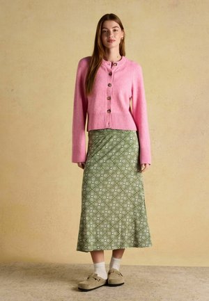 Frau steht und trägt eine rosa Knopfweste, einen grünen gemusterten Midirock, weiße Socken und beigefarbene Slipper vor einem beigen Hintergrund.