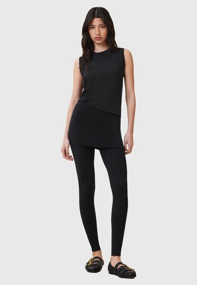 Haut noir sans manches associé à une mini-jupe à ourlet arrondi et des leggings noirs. Le mannequin porte des mocassins noirs avec des accents dorés.