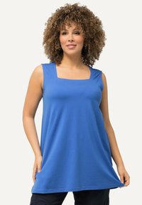 Donna con capelli ricci, che indossa un top senza maniche di colore blu e pantaloni neri, tenendo l'orlo del top con entrambe le mani.