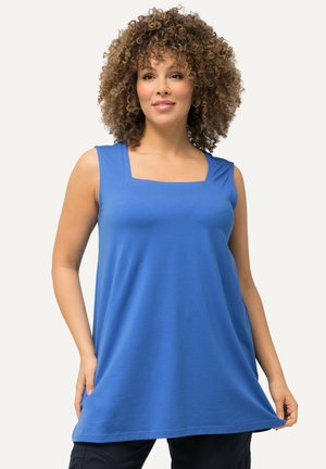 Vrouw met krullend haar, gekleed in een mouwloze blauwe tuniek en zwarte broek, terwijl ze de zoom van de tuniek met beide handen vasthoudt.