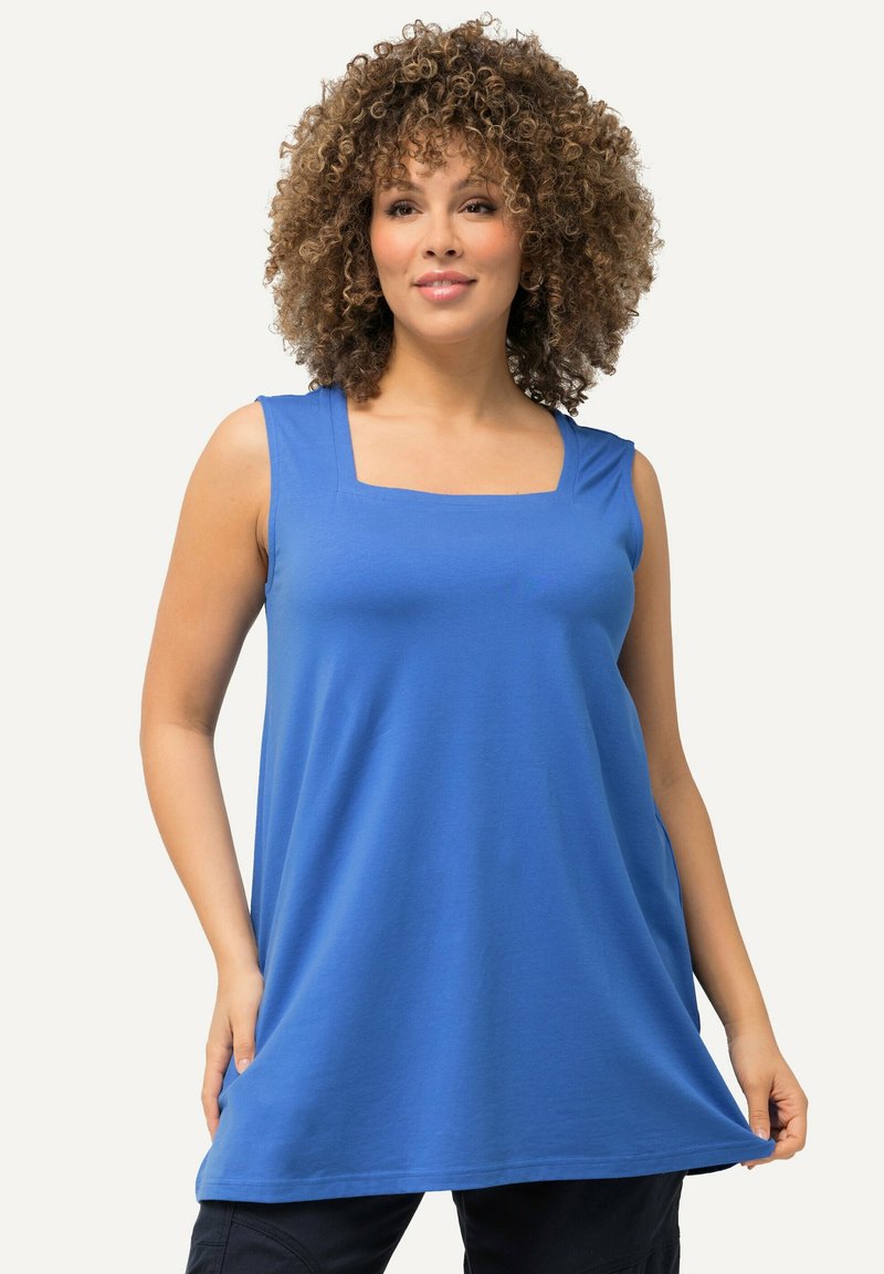 Donna con capelli ricci, che indossa un top senza maniche di colore blu e pantaloni neri, tenendo l'orlo del top con entrambe le mani.