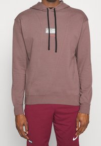 Hoodie bordeaux avec une poche avant, des cordons de serrage noirs et un patch logo réfléchissant. Dispose d'un tissu doux et de détails en panneaux sur les côtés.