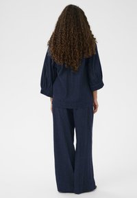 Sieviete ar gariem cirtainiem matiem, tērpusies brīvā tumši zilā garajās piedurknēs blūzē un platiem biksēm, stāv ar muguru pret skatītāju gaismas fonā.