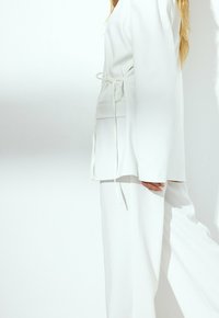 Costume blanc ajusté avec une coupe décontractée et une ceinture à la taille. Le tissu a l'air lisse avec un léger éclat, associé à un pantalon à jambes larges.