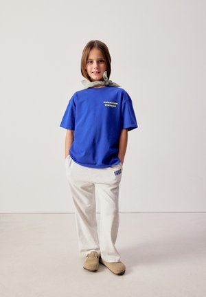 Mladé dievča stojí s rukami v vreckách, oblečené v modrom tričku s nápisom "American Vintage", bielych nohaviciach, béžových topánkach a šatke okolo krku.