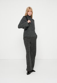 Suéter cinza tricotado com um decote alto e mangas longas, combinado com calças cinza texturizadas a condizer. O modelo segura uma pequena bolsa preta.