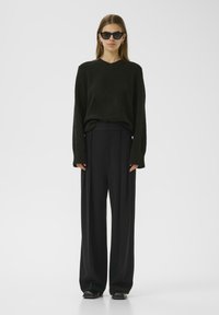 Black oversized sweater in textured fabric, paired with high-waisted black wide-leg trousers. Accessories include black sunglasses and earrings.