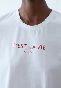 Bílý bavlněný tričko s červeným nápisem "C'EST LA VIE PARIS." Jednoduchý design s kulatým výstřihem a měkkou texturou.