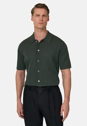 Mann trägt ein dunkelgrünes, kurzärmliges Hemd mit Knöpfen und schwarze Hose mit Stoffgürtel, steht vor einem einfarbigen Hintergrund.