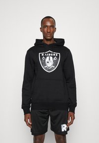 Sudadera negra con el logo de los Raiders, con un bolsillo canguro, capucha con cordón y puños acanalados. Llevada con pantalones cortos negros que presentan un logo blanco.