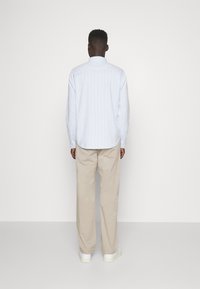 Jasnoniebieska koszula z długim rękawem w pionowe białe paski, z kołnierzykiem i guzikami przy mankietach, w połączeniu z beżowymi spodniami i białymi sneakersami. Widok z tyłu.