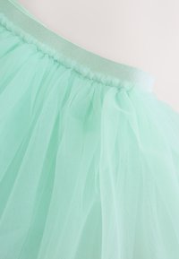 Jupe en tulle turquoise clair avec tissu transparent superposé et une ceinture élastique brillante sur un fond uni.