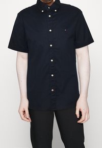 Camicia a maniche corte navy con colletto a punta, dotata di bottoni dorati e un piccolo dettaglio del logo sul petto. Texture liscia.