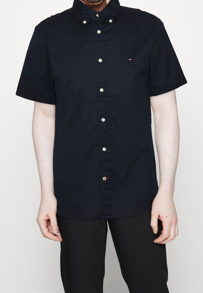Camicia a maniche corte navy con colletto a punta, dotata di bottoni dorati e un piccolo dettaglio del logo sul petto. Texture liscia.