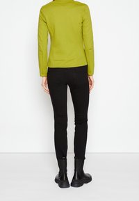 Veste cintrée verte à manches longues, associée à un jean noir skinny et des bottines noires, vue de dos. Texture lisse.