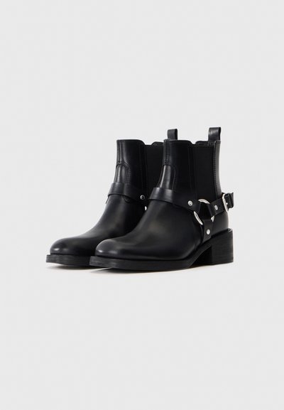 Botines de piel negra con correas de hebilla plateada, puntas redondeadas y tacones bajos tipo bloque sobre un fondo claro y liso.