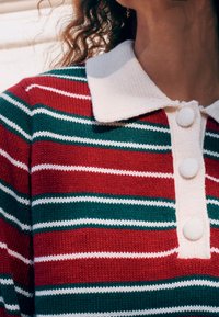 Pull tricot rayé rouge et vert avec un col crème et trois boutons texturés à l'avant.