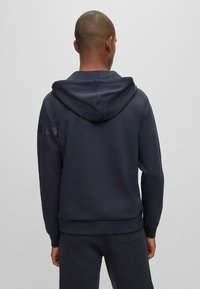 Mörk marinblå huvtröja i slätt tyg, med en kenguruficka och ribbade ärmslut, samt diskreta randiga detaljer på axeln.