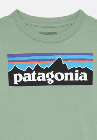 Zeleni pamučni majica s crnim pravokutnikom na kojem stoji riječ "patagonia" bijelom bojom, iznad grafike višebojne planine.