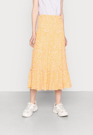 Person, die einen gelb-weißen gemusterten Midirock, weiße Sneakers und ein in die Hose gestecktes helllila Oberteil trägt, vor einem schlichten Hintergrund steht.