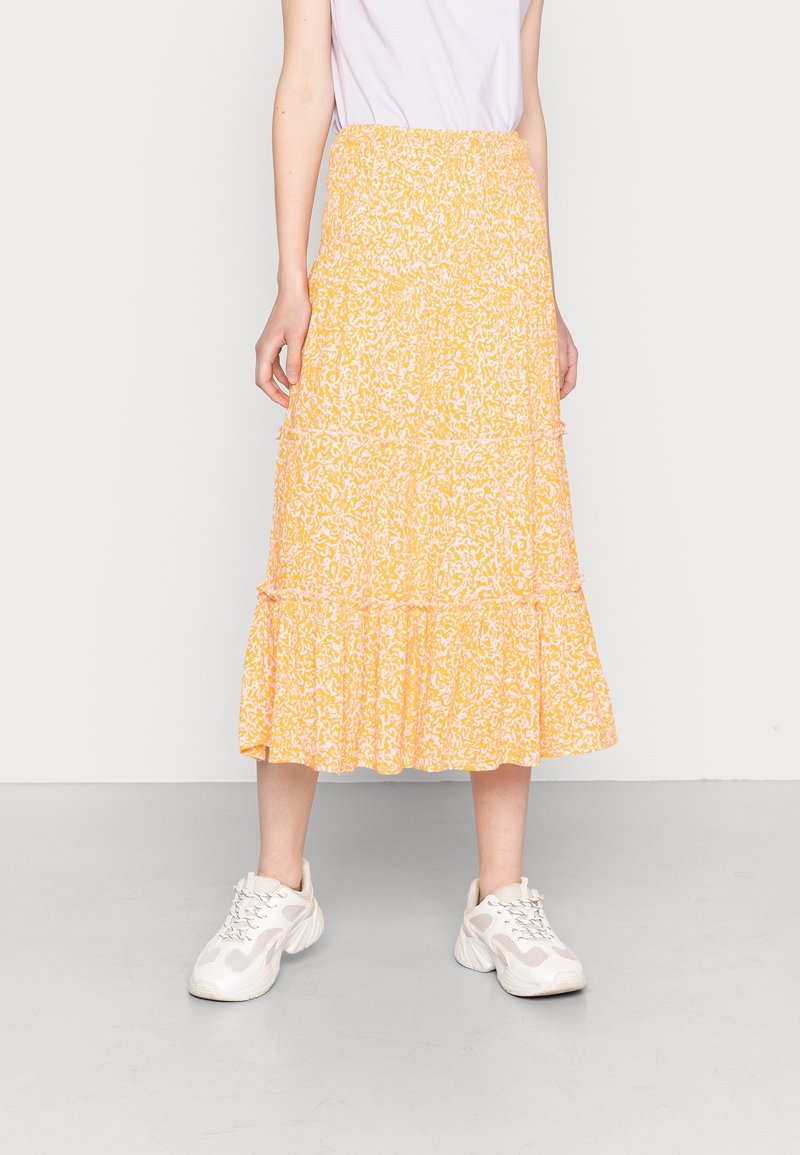 Henkilö, joka pukee ylös keltaisen ja valkoisen kuvioidun midihameen, valkoiset sneakers-kengät ja keveän violetti paidan, seisoo tasaisessa taustassa.