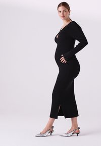 Čierne pletené šaty so žebrovaným vzorom, hlbokým výstrihom do V, dlhými rukávmi a bočnými rozparkami. Kombinované s metalickými striebornými lodičkami na opätku s remienkom a minimalistickými detailmi.
