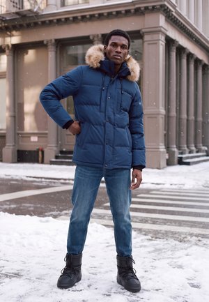 Homme debout dans une rue de la ville enneigée portant un manteau d’hiver bleu avec capuche en fourrure, un jean et des bottes d’hiver noires.