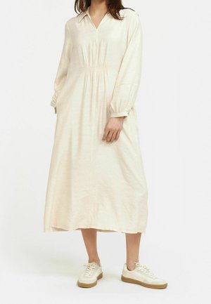 Abito lungo color crema con colletto e vita arricciata, dotato di maniche lunghe e una superficie liscia. Abbinato a sneakers bianche.