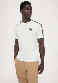 White short-sleeve T-shirt with black shoulder stripes and a small EA7 logo patch on the front. Smooth texture and classic round neck design.