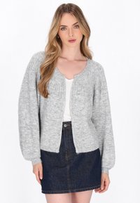 Tânără cu păr lung blond, îmbrăcată cu un cardigan tricotat gri deschis peste un top alb și o fustă din denim închis, stând în fața unui fundal alb.
