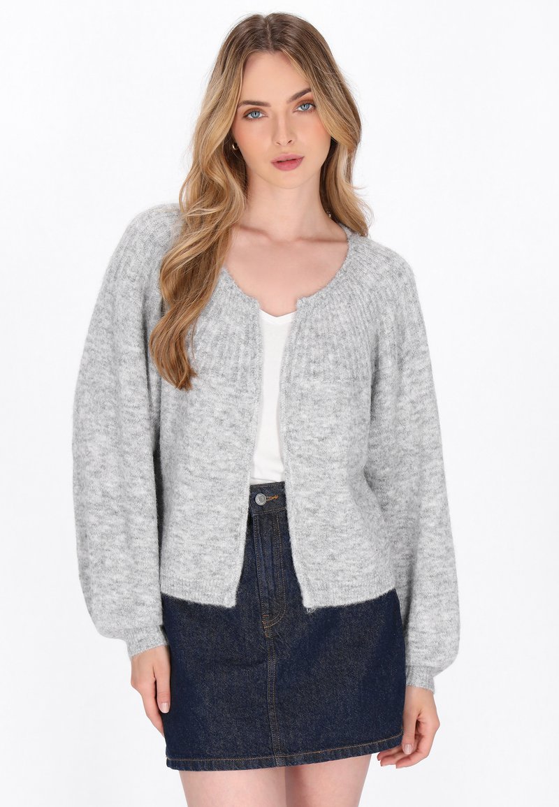 Tânără cu păr lung blond, îmbrăcată cu un cardigan tricotat gri deschis peste un top alb și o fustă din denim închis, stând în fața unui fundal alb.